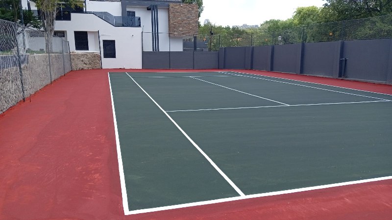 Newly built outdoor tennis court with green playing surface and red border, surrounded by high fencing next to a modern residential building.